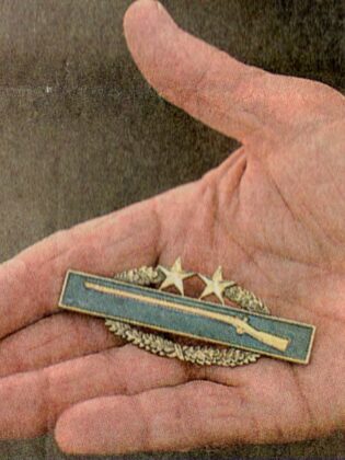 John McCrimmon's hand with a combat award, depicted as a rifle with a laurel wreath, and two stars on top